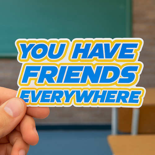 Hand holding a sticker with 'YOU HAVE FRIENDS EVERYWHERE' text in front of a blurred classroom background.