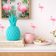 Decorative setup with pineapple, books, and flowers on a shelf against a wall with flamingo decals.